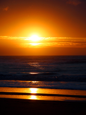 [The sun is a gold orb in the sky with an orange ring around it. The sun backlights the horizontal clouds as was as a strip of ocean and turns the wet sand gold.]